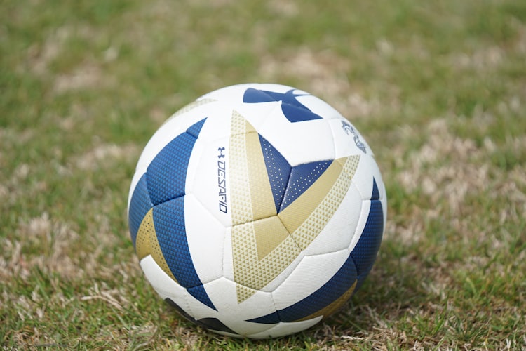 Bola sepak berwarna putih, biru, dan emas tergeletak di atas rumput lapangan